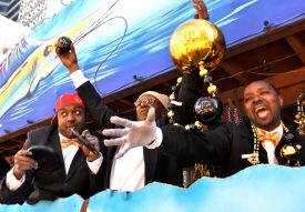 Zulu float riders, one of whom is holding up a coconut in one hand and in the other, a strand of Mardi Gras beads strung with a gigantic gold ball