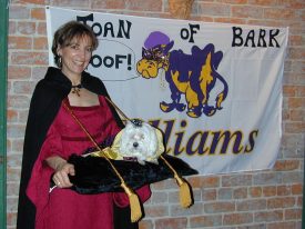 The white Maltese — seated on a black velvet pillow supported by a tray and a rope that wraps around the back of Karen’s neck — looks positively regal in her purple-and-gold cape.